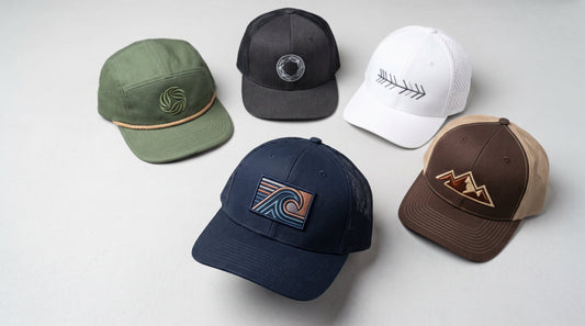 Five embroidered baseball caps in different styles and colors arranged on a table, each with a unique abstract stitched emblem, showing the quality and variety a custom hat embroidery decorator can produce